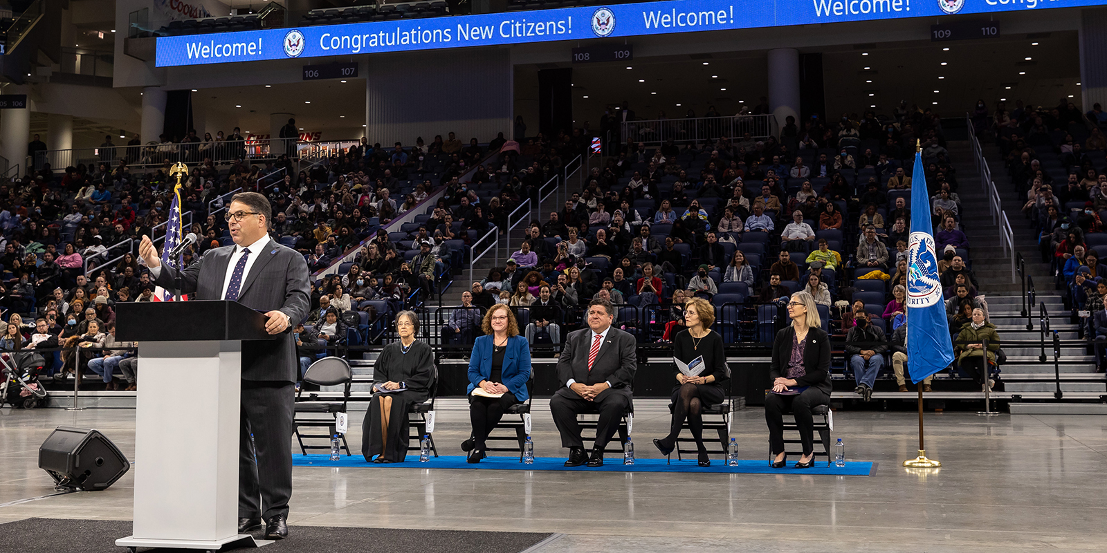 President Rob Manuel stands at a podium in the center of an arena floor, behind him sits dignitaries. 