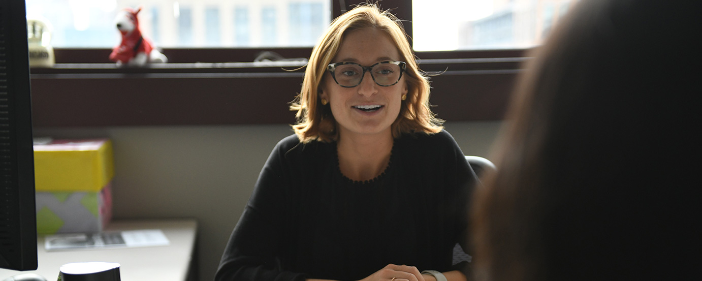 Woman wearing glasses, seated behind work desk, talking