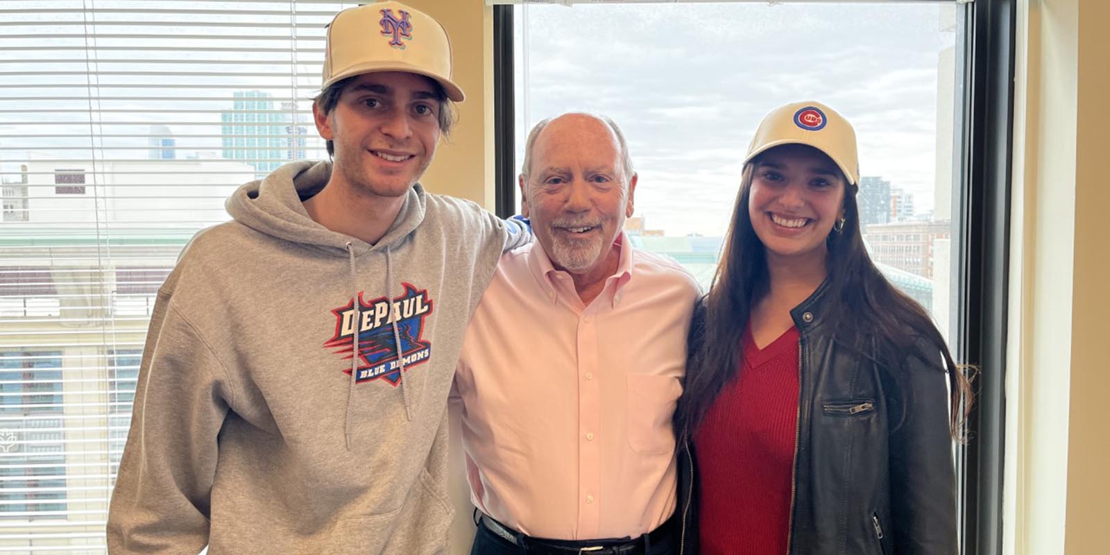 Two students, with baseball caps on, stand with their public relations professor in a classroom