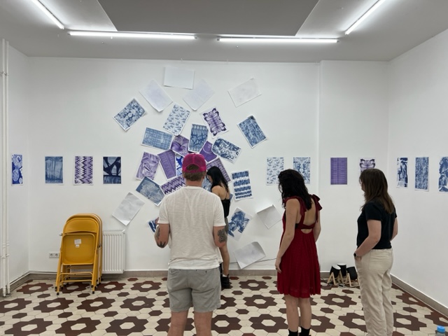 Students are standing with their back facing the camera toward a wall with many blue and purple abstract risograph prints in a cluster on the wall. More prints create a straight line across the other walls.