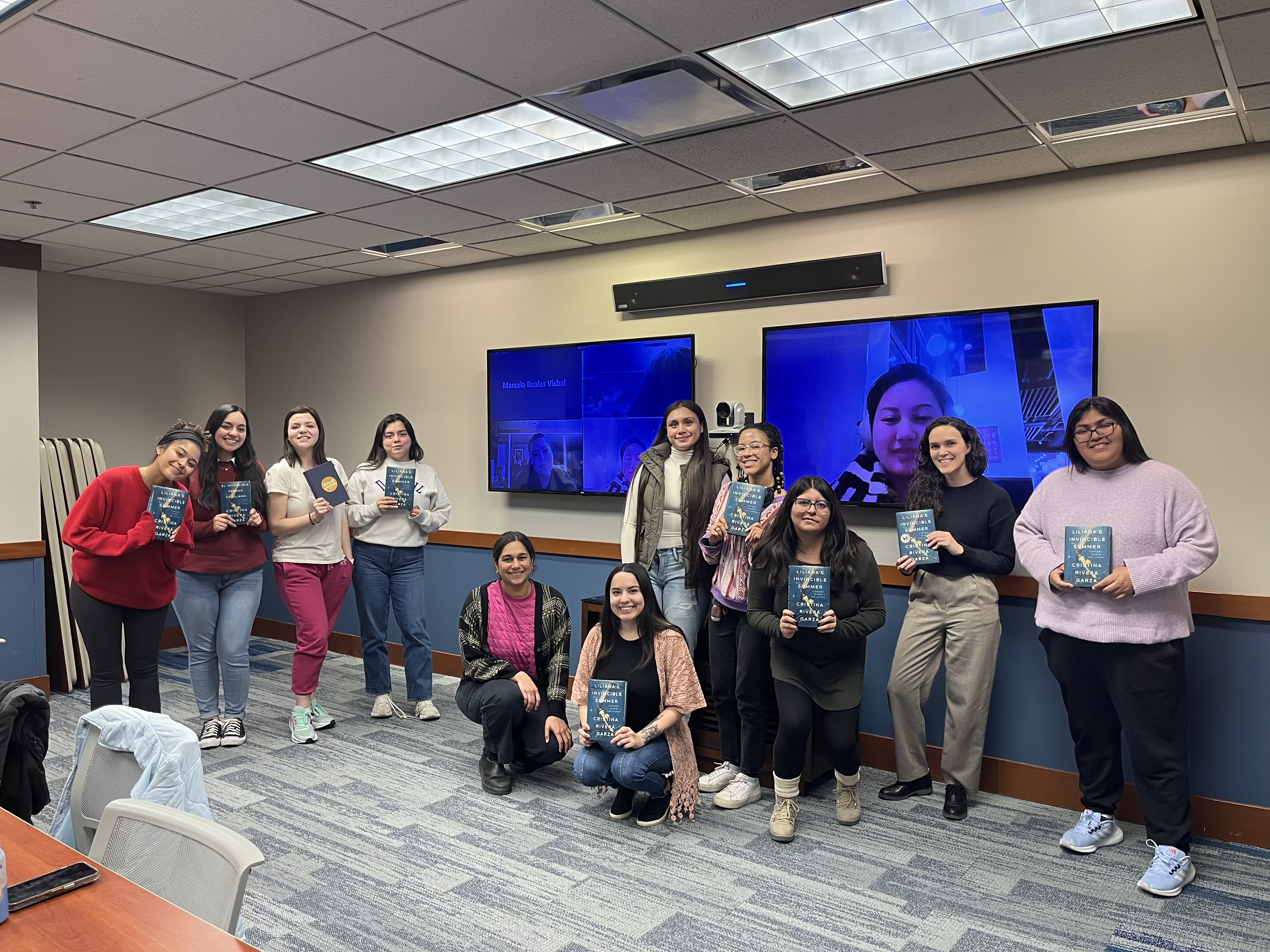 Latinx Book Club members holding their book and taking a picture including members joining via zoom in a meeting room at the DePaul Center
