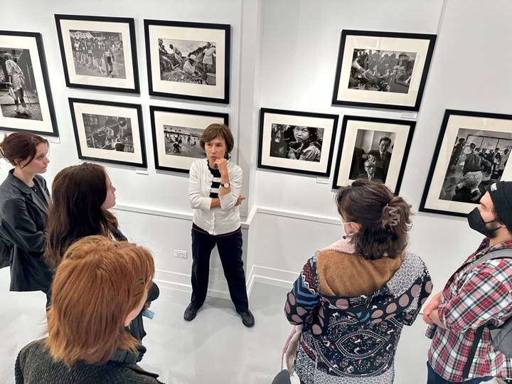 Aileen Mioko Smith standing in front of a wall with many black and white photographs, talking to a group of students.