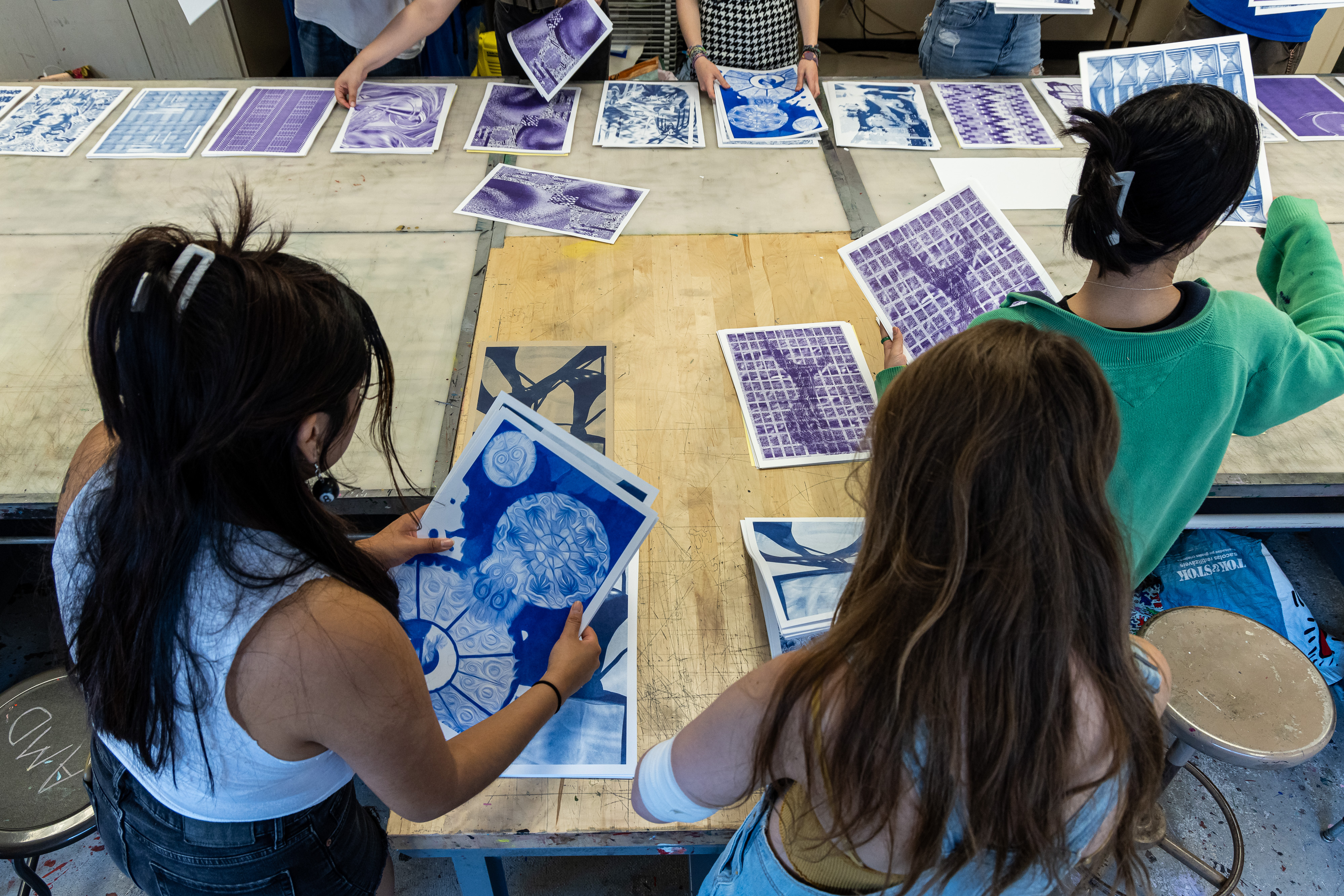 A photo taken from behind three students as they hold up a few of the abstract prints on the table