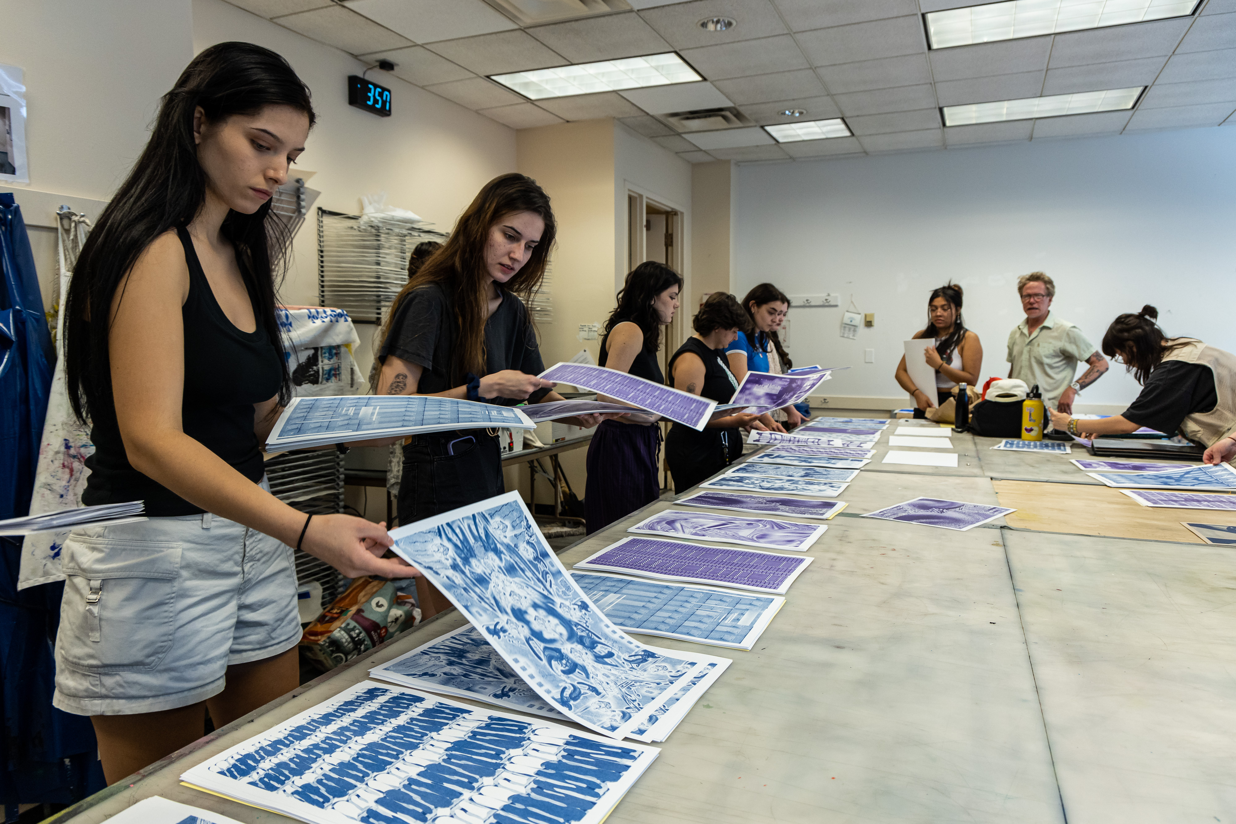 Many students are lined up along a table where they pick up many different prints that are on the table.