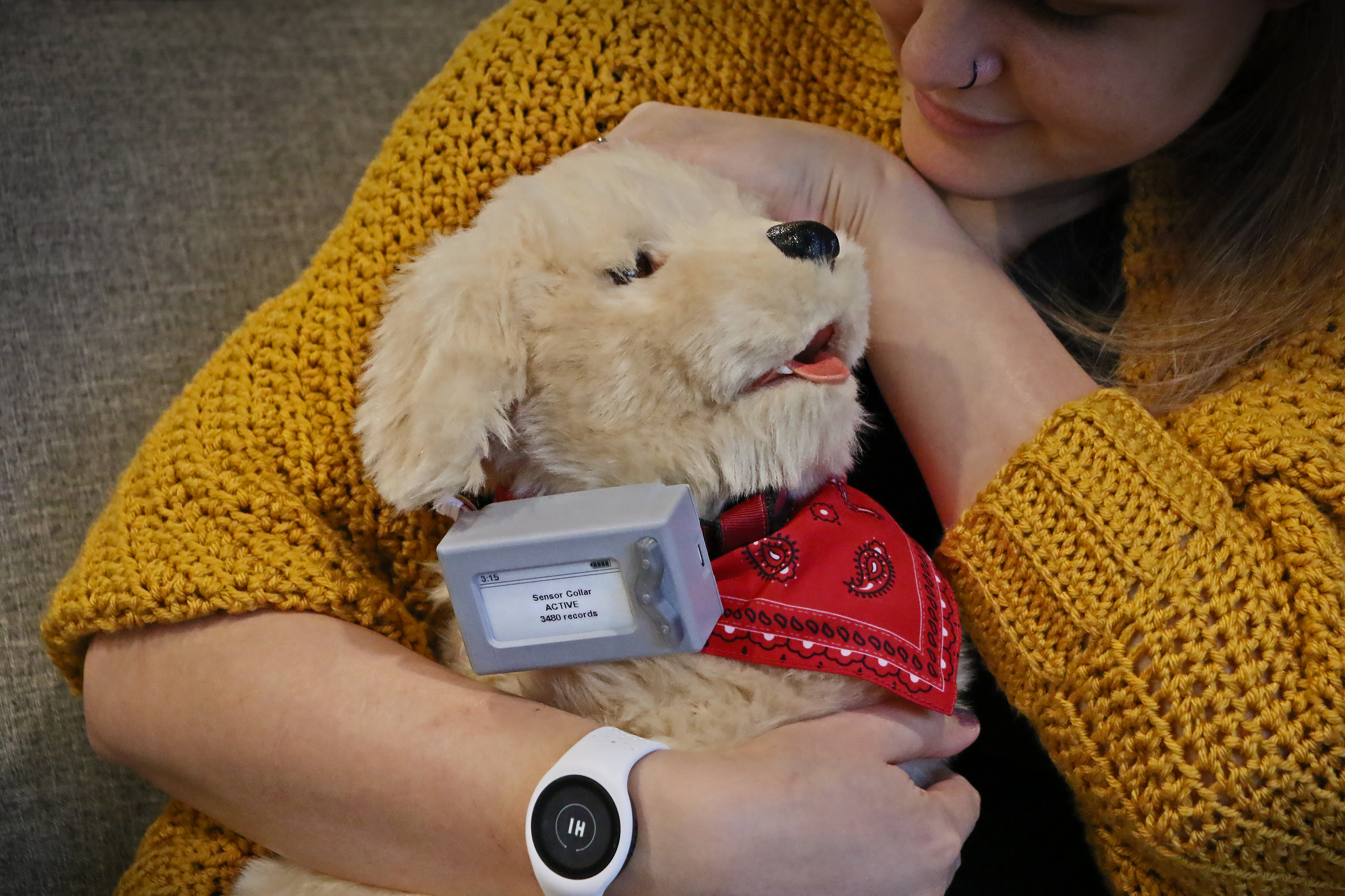 A woman wearing a yellow crocheted cardigan holds a robot dog with a wristband on her wrist.