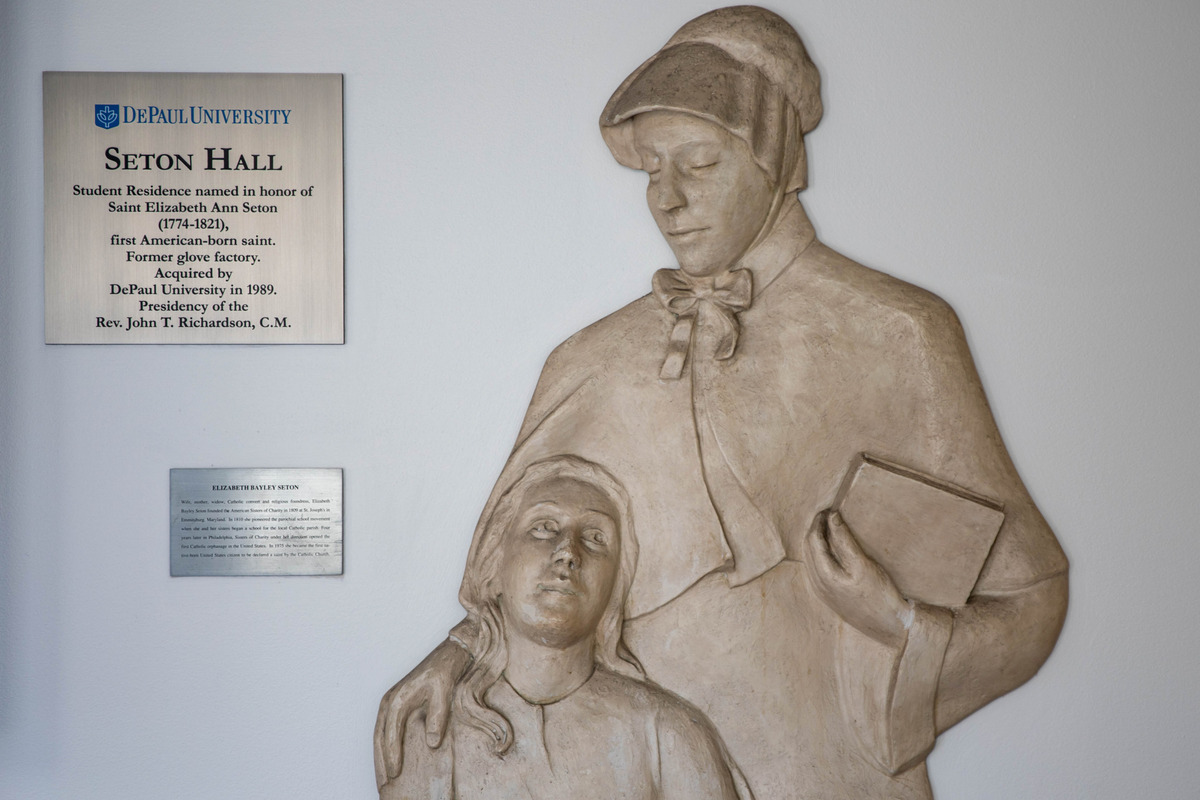 A wall statue of Saint Elizabeth Ann Seton holding a book with her arm around a girl, there is a plaque explaining that the student residence, Seton Hall, was named after her.