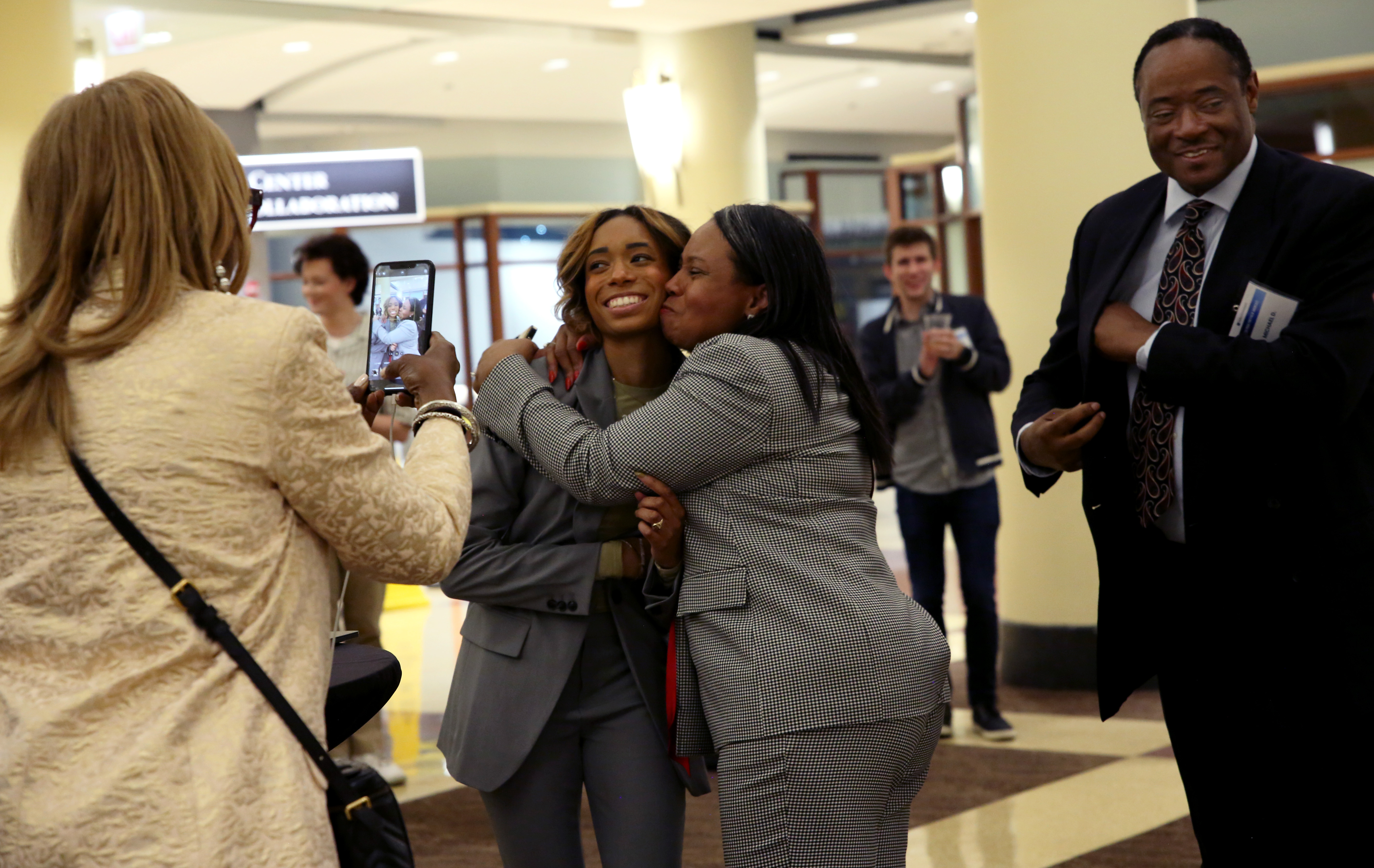 Woman hugs student and kisses her cheek as someone takes their photo on a phone.