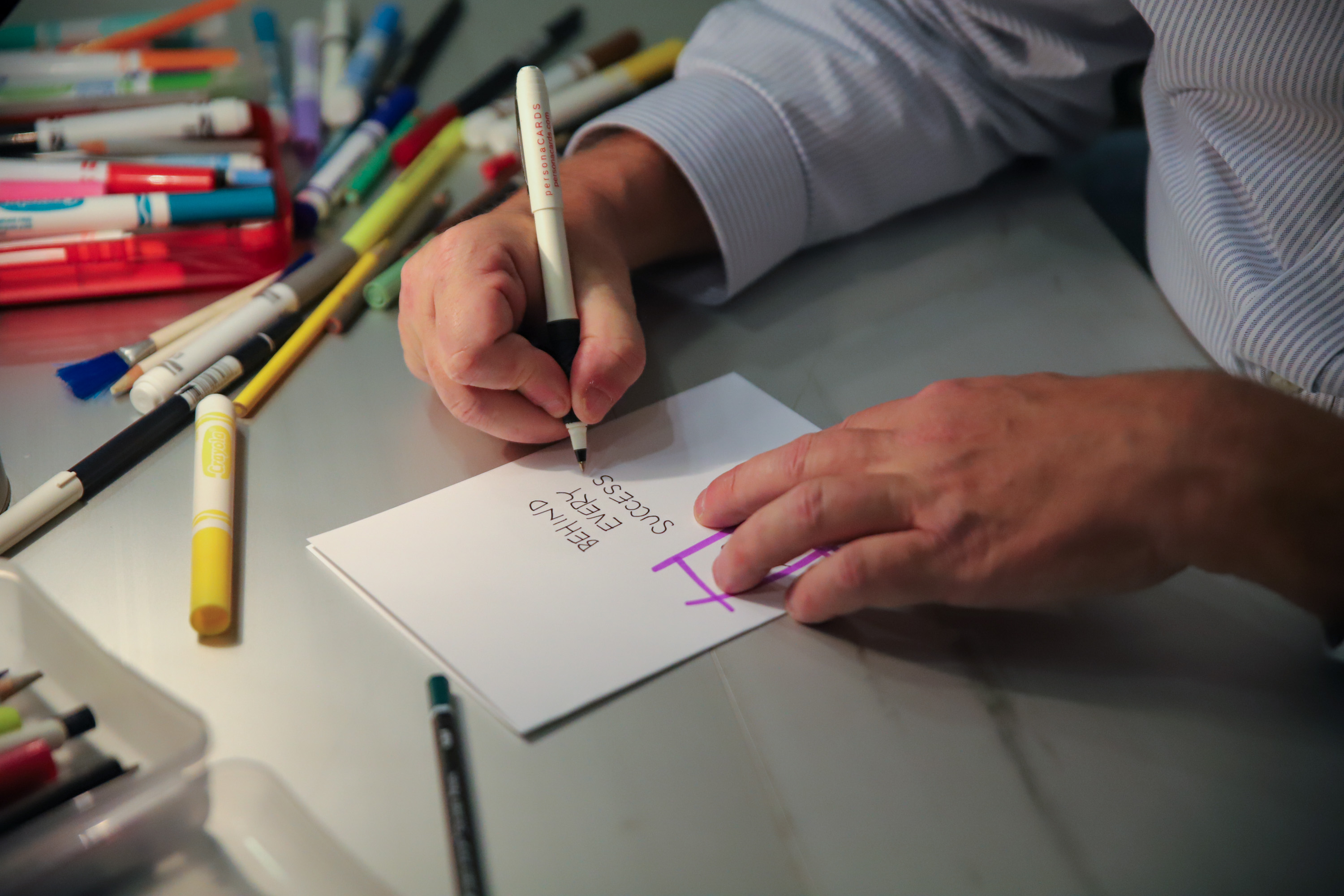 Man writes 'Behind every success' on a card with markers and pencils on the table next to him.