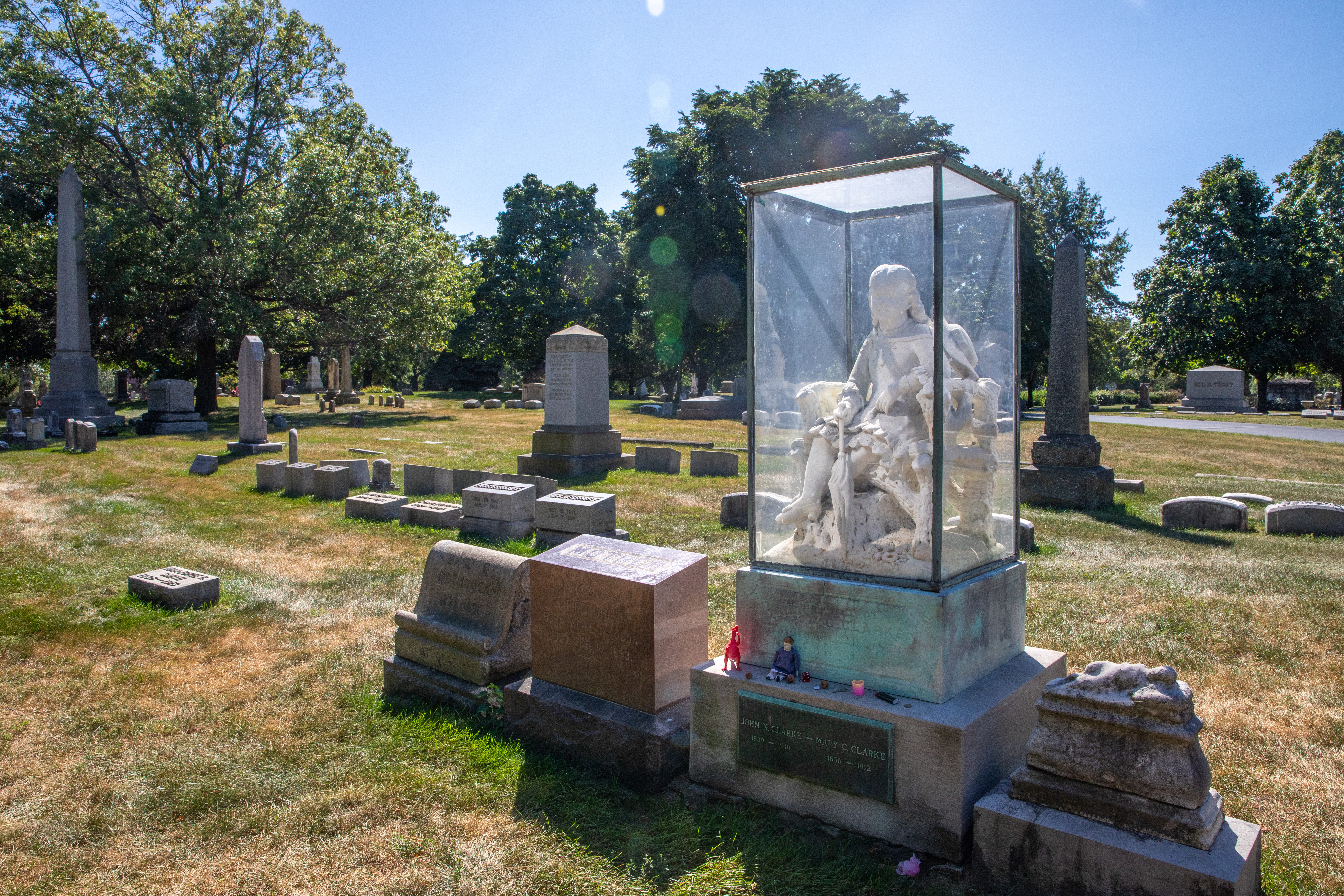 Inez Briggs' grave in Graceland Cemetery, surrounded by greenery and other graves around her. Her grave includes a statue of a little girl encased in a glass box. There are toys and small trinkets left near the bottom of the grave.