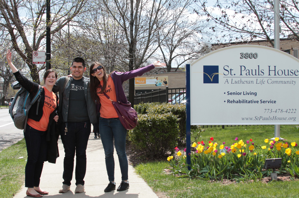 Three students smiling and posing in front of the St. Pauls House senior living building.